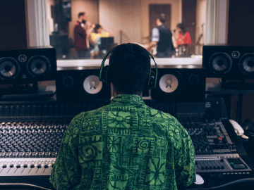 Male sound engineer sits at a recording desk with headphones on. Musicians are visible in the recording booth