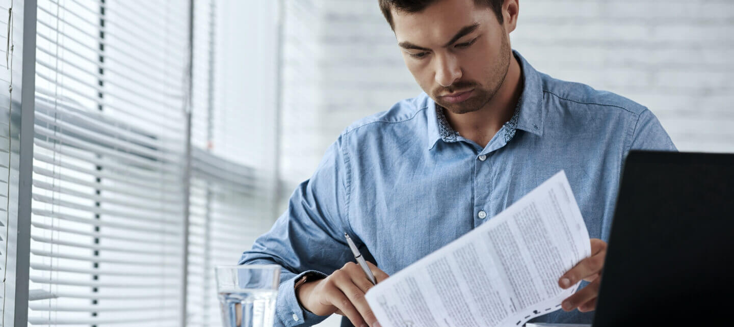 A serious contractor in a blue shirt reviewing tax documents at a desk with a laptop and a glass of water, symbolising IR35 compliance and financial scrutiny.
