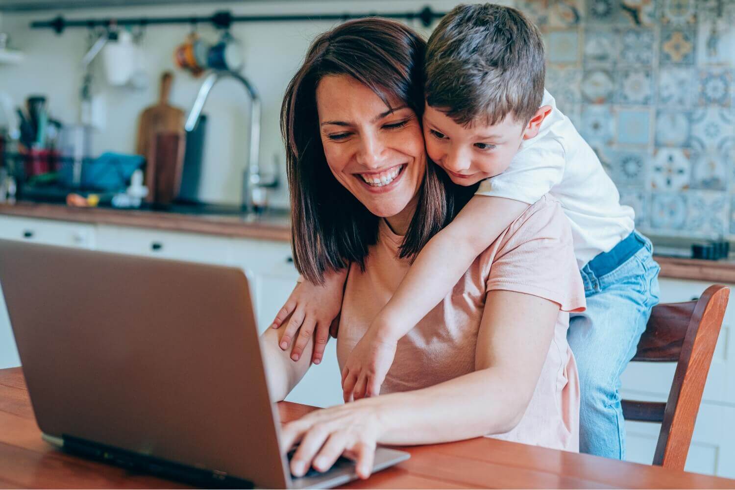 Freelancer mother smiling at laptop while child hugs her from behind in home kitchen, representing work-life balance and family time